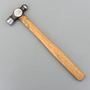 以下が含まれることがあります： 木製の柄が付いたボールピンハンマー。 ハンマーヘッドは銀と黒で、片側に丸みを帯びた打撃面、もう片側にボール型のピンが付いています。 柄は明るい色の木材で作られています。