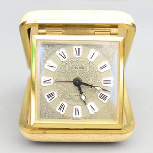 以下が含まれることがあります： 金色で正方形の目覚まし時計で、白い文字盤に黒いローマ数字の時間の目盛りがあります。時計の文字盤には、装飾的な花の模様と「EUROPA」と「2 JEWE」の文字が印刷されています。この時計はドイツ製です。