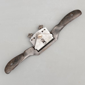 May include: A vintage drawknife with a metal blade and two textured handles. The blade is held in place by two adjustment screws. The tool is made of metal and has a worn appearance, suggesting it is an antique woodworking tool.