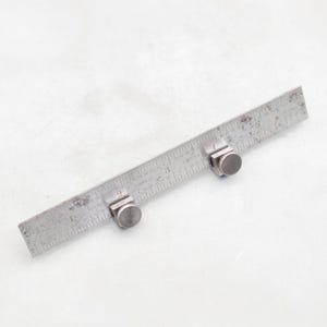 May include: A vintage metal ruler with measurements in inches. The ruler has a worn, aged appearance and is marked with numbers and lines. Two adjustable metal stops are attached to the ruler, adding functionality. The tool is designed for precise measurements.