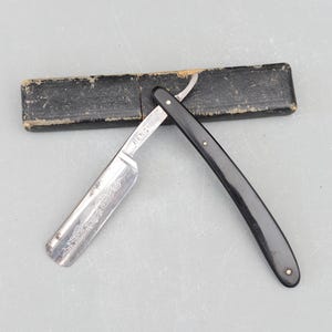 Puede incluir: Una navaja de afeitar abierta con mango negro y hoja plateada, junto a su estuche de cuero negro. La hoja está marcada con la palabra "BENGAL" y grabados decorativos.