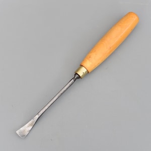 Puede incluir: Una gubia para trabajar la madera con una hoja curva de color plateado y un mango de madera clara. El mango es cilíndrico y está unido a la hoja con una virola de latón. La herramienta está diseñada para tallar y dar forma a la madera.