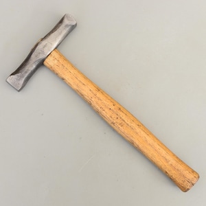 May include: A metal hammer with a wooden handle. The hammer head is a dark gray color and the handle is a light brown color. The hammer is angled to the right.