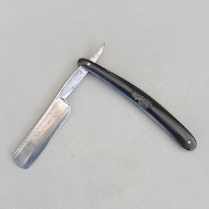 May include: A vintage straight razor with a black plastic handle and a silver blade. The blade is engraved with the text "MAX SCHWADOP & SOHN SOLINGEN".
