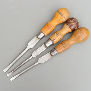 Może przedstawiać: Trzy vintage śrubokręty z drewnianymi uchwytami. Uchwyty są jasnobrązowe, a metalowe ostrza są srebrne. Śrubokręty są ułożone w kształcie trójkąta.