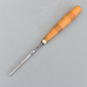 May include: A woodworking chisel with a light brown wooden handle featuring horizontal grooves. The metal blade is silver and tapers to a flat end. A brass ferrule connects the handle to the blade. The tool is designed for shaping wood.