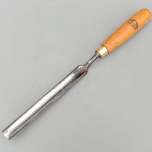 Puede incluir: Un formón para carpintería con mango de madera y hoja de metal. El mango es de color marrón claro con una virola de latón. La hoja es plateada y se estrecha hasta un filo. El formón está diseñado para dar forma a la madera.