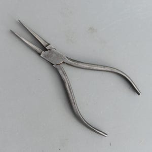 Può includere: Una pinza a becchi lunghi, un utensile manuale con ganasce lunghe e affusolate per afferrare piccoli oggetti. La pinza è in metallo e ha manici curvi. Progettata per lavori di precisione, è comunemente utilizzata nella gioielleria e nell'elettronica.