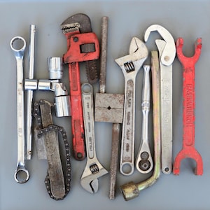 Puede incluir: Un surtido de herramientas manuales, que incluyen llaves, vasos y una llave de cadena, dispuestas sobre una superficie gris. Las herramientas son una mezcla de plata y rojo, mostrando signos de uso. La imagen muestra varias herramientas para trabajos mecánicos.