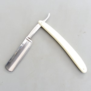 Peut inclure: Un rasoir droit vintage avec un manche en plastique blanc. Le rasoir a une lame argentée et un petit manche incurvé.