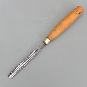 May include: A woodworking chisel with a polished steel blade and a light brown wooden handle. The handle is cylindrical and tapers slightly towards the end. A brass ferrule connects the blade and handle. The chisel is designed for shaping wood.