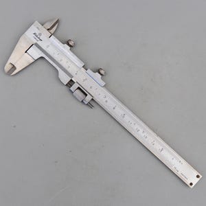 May include: A silver Mitutoyo caliper, a precision measuring tool, is shown on a grey surface. The caliper has a sliding scale with both inch and centimetre markings, and a small adjustment screw. The tool is designed for accurate measurements.