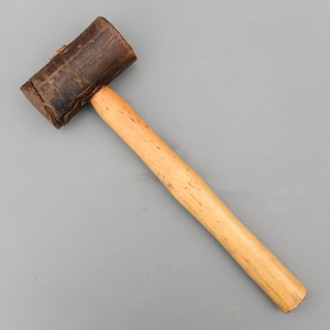 May include: A mallet featuring a cylindrical head wrapped in dark brown textured material and a smooth, light-colored wooden handle. The tool has a straight, cylindrical shaft and a head that is securely attached to the top of the handle.