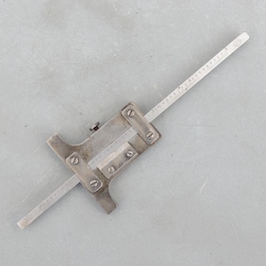 May include: A vintage metal depth gauge with a sliding scale and measurement markings. The gauge is silver-toned with a rectangular body and a long, thin measuring arm. The scale is marked in inches.