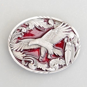 Peut inclure: Boucle de ceinture en argent avec un fond en émail rouge représentant un aigle en vol avec des plumes et des détails ornés.