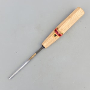 May include: A wood carving chisel with a light-colored wooden handle and a metal blade. The handle has a red and gold label with the word "TIRANT" printed on it. The chisel is designed for detailed woodworking.