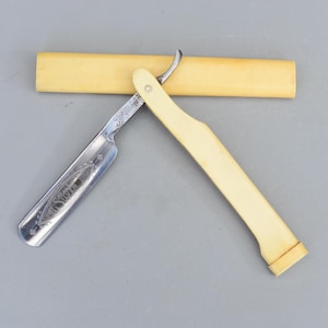 Puede incluir: Una navaja de afeitar recta antigua y abierta con un mango color crema y una hoja plateada. La hoja está grabada con la palabra "Silver" y diseños florales. Detrás de la navaja hay una funda color crema.