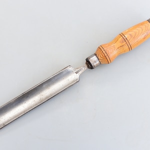 May include: A woodworking chisel with a light brown wooden handle and a silver metal blade. The handle has a rounded shape with decorative grooves. The blade is long and rectangular, with a slightly curved cutting edge. The tool is designed for shaping wood.