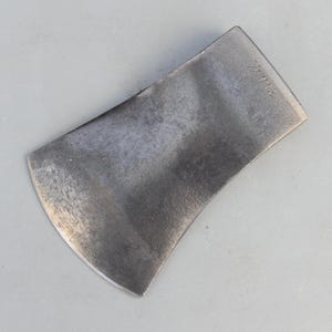 May include: A dark grey metal axe head with a curved cutting edge and a flat poll. The surface of the metal shows a weathered, textured finish with visible signs of use. The word "TRUE" is stamped into the metal near the top corner of the head.