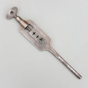 Può includere: Una chiave per maschi in metallo vintage con un lungo manico e una testa a forma di chiave. La chiave ha un corpo rettangolare con una serie di fori numerati e un meccanismo a vite per tenere i maschi. L'utensile è in metallo scuro e invecchiato.