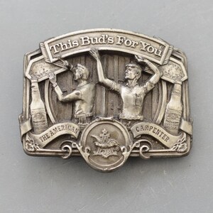 Peut inclure: Boucle de ceinture en métal argenté avec le texte "This Bud's For You". La boucle présente deux personnages, des bouteilles de bière et les mots "The American Carpenter". Le logo Budweiser est également visible.