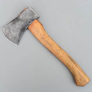 Puede incluir: Un hacha pequeña con un mango de madera y una cabeza de acero. La cabeza del hacha es de color gris oscuro y el mango de madera es de color marrón claro. El hacha está diseñada para cortar madera y otras tareas.