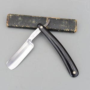 以下が含まれることがあります： 光沢のある銀色の刃と黒い柄のストレートカミソリ。カミソリは開いており、鋭い刃先を示しています。使い古された黒いケースがカミソリの後ろにあります。