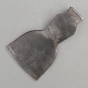 May include: A worn, dark grey axe head with a curved blade and a flared, rectangular cutting edge. The metal shows signs of wear and use, with a slightly textured surface. The axe head is designed for woodworking or similar tasks.