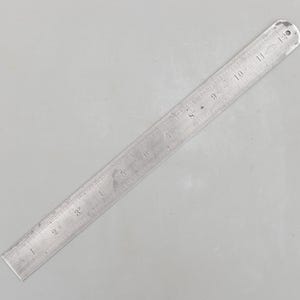 Peut inclure: Une règle métallique vintage avec des marquages gravés, numérotée de 1 à 30 cm. La règle est de couleur argentée avec un petit trou à l'extrémité de 30 cm. Les marquages sont clairement visibles.