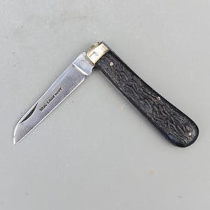 May include: A partially open pocket knife with a silver blade and a black textured handle. The blade has the text "W.R. Case & Sons" etched on it. The knife has a brass bolster and rivets.