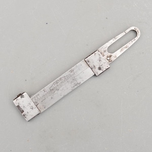 Op de afbeelding: Een vintage metalen liniaal met een schuifmechanisme en een lus aan één uiteinde. De liniaal heeft geëtste markeringen voor inches en fracties van een inch. De tekst "CHESTERMAN SHEFFIELD ENGLAND" is zichtbaar. De liniaal wordt waarschijnlijk gebruikt om te meten.