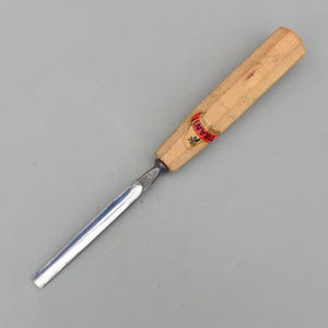 以下が含まれることがあります： 明るい色の木製の柄と銀色の刃が付いた木彫りのノミ。ハンドルには「IRANI」の文字が印刷された赤と金のラベルが付いています。ノミは灰色の背景に対して斜めに配置されています。