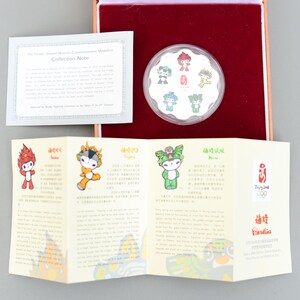 以下が含まれることがあります： 北京2008年オリンピックのマスコット「フレンドリーズ」をデザインした銀製の記念硬貨5枚セット。赤いベルベットの箱に、中国語と英語でマスコットの名前が書かれた白いカードと共に展示されています。