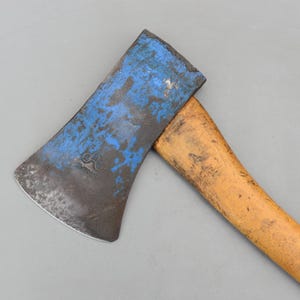 Könnte beinhalten: Ein Axtkopf aus Metall mit abgenutzter blauer Lackierung und einem sichtbaren eingestanzten Logo, befestigt an einem hellbraunen Holzstiel. Die Metallklinge weist Gebrauchsspuren mit dunkler Oxidation und Kratzern auf, der Stiel ist glatt.