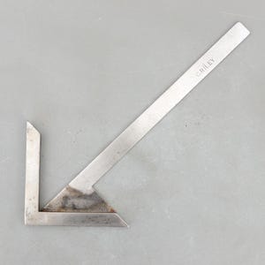 May include: A vintage metal try square with a right angle. The tool has a long, thin arm with the text "C. RILEY" and a shorter arm. The metal has some discoloration, suggesting age and use. The try square is used for woodworking and carpentry.
