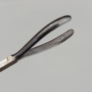 May include: Close-up of a metal tool with curved arms. The tool is dark gray with the word "VANALOY" etched on one arm. The tool's design suggests it is used for gripping or manipulating small objects.