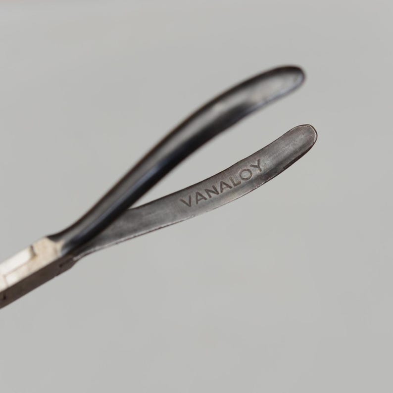 May include: Close-up of a metal tool with curved arms. The tool is dark gray with the word "VANALOY" etched on one arm. The tool's design suggests it is used for gripping or manipulating small objects.