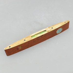 May include: A vintage wooden level with brass accents. The level features a clear vial filled with green liquid to indicate levelness. A small, round, blue and white sticker is affixed to the side. The level is approximately 12 inches long.