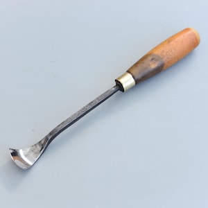 May include: A woodworking gouge with a curved blade and a wooden handle. The handle is light brown with a dark brown section near the metal ferrule. The blade is curved and made of steel. The tool is designed for carving.
