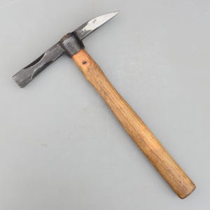 Puede incluir: Un pico martillo vintage con mango de madera y cabeza de metal. La cabeza tiene un extremo puntiagudo y otro plano. El mango es de madera marrón claro. El martillo está diseñado para romper rocas u otros materiales duros.