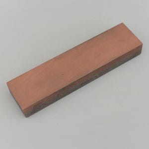 May include: A rectangular sharpening stone with a reddish-brown top surface and a dark grey bottom edge. The stone is designed for sharpening knives and other cutting tools. The stone is approximately 18 cm long.