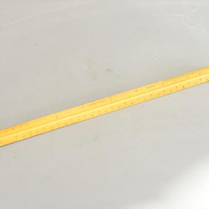 May include: A yellow architectural scale ruler with markings and measurements. The ruler is made of a light-colored material and is used for drafting and design work. It is approximately 12 inches long.