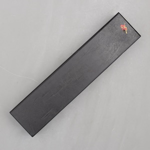 May include: A long, rectangular, black wooden box with a small, red and gold decorative element on the top right corner. The box has a slightly textured surface and is angled diagonally.