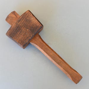 Könnte beinhalten: Ein Holzhammer mit braunem Stiel und einem rechteckigen Kopf. Der Kopf ist aus dunklem Holz mit einer Maserung.