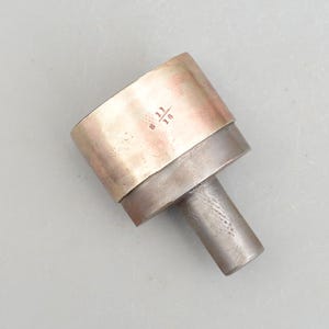 Op de afbeelding: Een metalen gereedschap met een messingkleurige bovenkant en een zilverkleurige onderkant. De bovenkant heeft de nummers "6 11 18" gegraveerd.