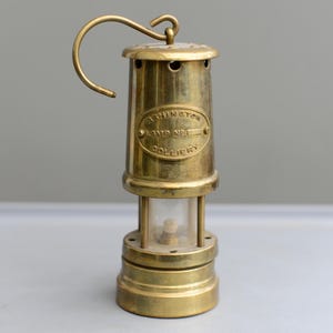 May include: Antique brass miner's lamp with a curved handle. The lamp has a glass enclosure and an oval brass plaque with the text "ASHTON LAMP NO. 1 COLLIERY". The lamp is cylindrical and made of brass.
