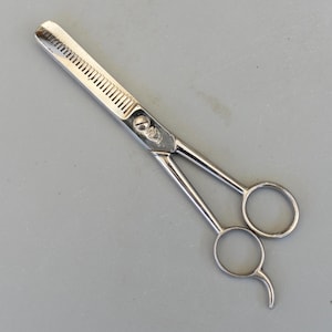 Puede incluir: Un par de tijeras plateadas para adelgazar el cabello. Una hoja tiene dientes muy juntos, mientras que la otra es una hoja sólida. Las tijeras tienen anillos para los dedos para facilitar el manejo. Las tijeras están diseñadas para peluquería profesional.