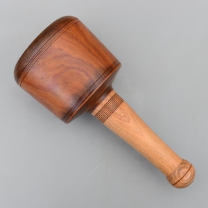 Puede incluir: Un mazo de madera con una cabeza marrón oscuro y un mango marrón claro. La cabeza tiene dos anillos tallados y el mango tiene un extremo redondeado. El mazo se utiliza probablemente para la carpintería o la artesanía.