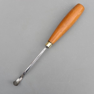 Puede incluir: Una herramienta de tallado de metal con mango de madera y un extremo curvado en forma de cuchara. El mango es de color marrón claro y el vástago metálico es plateado. La herramienta está diseñada para tallar y dar forma a madera.