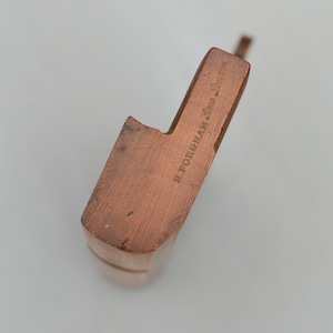 May include: A copper-coloured pipe with a rectangular head and a curved stem. The text "H. FORGHAM Lime London" is engraved on the side. The pipe is made of a solid material and has a smooth finish.